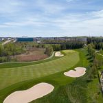 A golf course with several sand pits from a bird's eye view. Houses and commercial buildings are visible in the distance.