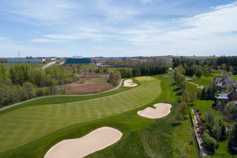 A golf course with several sand pits from a bird's eye view. Houses and commercial buildings are visible in the distance.