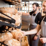 Two male bakery employees wearing gray aprons and handling loaves of bread. The bread is in baskets on the wall.