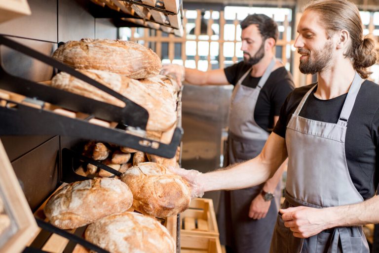 Two male bakery employees wearing gray aprons and handling loaves of bread. The bread is in baskets on the wall.