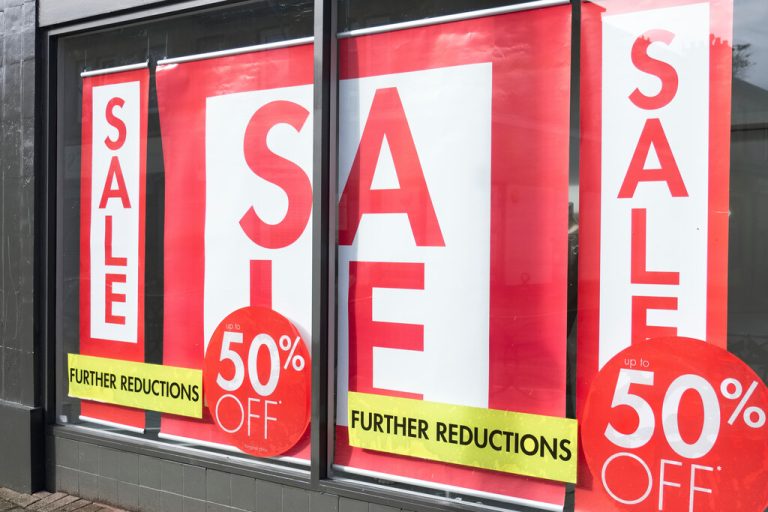 A storefront business with hanging window banners advertising a big sale inside the store. The signs are red and white.