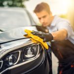 A man wearing a blue shirt and black gloves uses a yellow hand cloth to polish the exterior of a black vehicle.
