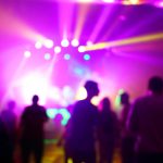 A crowd of people standing in front of a bright stage at an event. The stage is illuminated with purple lights.
