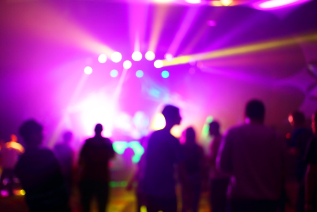 A crowd of people standing in front of a bright stage at an event. The stage is illuminated with purple lights.