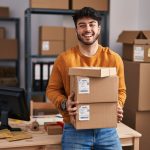 A young man wearing an orange sweater smiles widely as he holds three cardboard boxes in front of a desk.