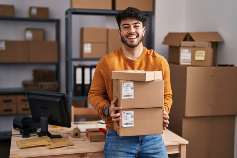 A young man wearing an orange sweater smiles widely as he holds three cardboard boxes in front of a desk.