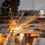 A large industrial cutting machine with sparks flying off of it operates on a sheet of metal in a workshop.