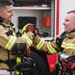 Two firefighters in their coats and safety gear as they smile and shake hands with one another. The truck is behind them.