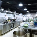 A commercial kitchen with chefs hard at work. The kitchen has stainless steel features and steam blowing off surfaces.