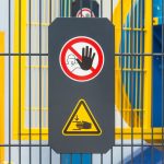 A sign on a metal fence guarding machinery. The sign displays safety symbols for safe machine operation.