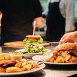 Four plates of food sit on a stainless steel table in a restaurant kitchen, with two staff members behind.