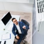 An overhead view of an air conditioner in a room, with a man in a suit sitting at a desk below, pointing a remote at the AC.