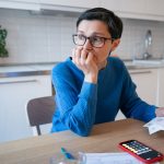 A stressed out middle-aged woman sitting at her dining room table holding her paycheck. She appears worried.