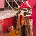 A worker wearing a red jump suit stands at the pack of a truck while tightening a latching strap securing cargo.