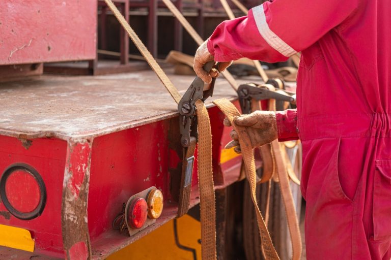 A worker wearing a red jump suit stands at the pack of a truck while tightening a latching strap securing cargo.