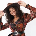 A young Black woman wears a red and black paisley-printed dress with a black belt and a brown cowboy hat.