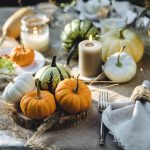 A fancy table contains a burlap overlay, small pumpkins, white plates, candles, cutlery, and wine glasses with white wine.