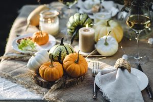 A fancy table contains a burlap overlay, small pumpkins, white plates, candles, cutlery, and wine glasses with white wine.