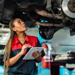 A mechanic stands underneath a lifted car in an automotive shop and inspects the undercarriage while holding a clipboard.