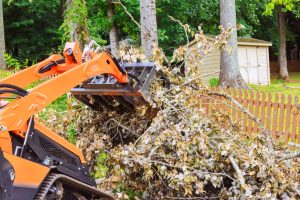 An orange skid steer with a gripping attachment removes leaves and branches of debris from a yard near a fence line.