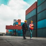 Two people wearing yellow hard hats, one carrying a clipboard, walk near shipping containers.
