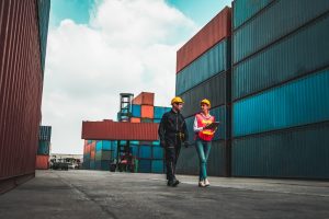 Two people wearing yellow hard hats, one carrying a clipboard, walk near shipping containers.