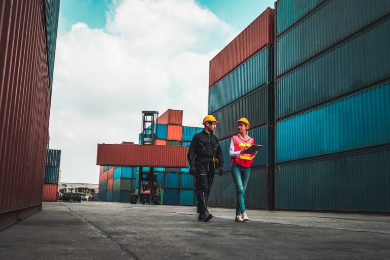 Two people wearing yellow hard hats, one carrying a clipboard, walk near shipping containers.