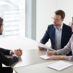 A man and woman taking notes and sitting across from a man at a table. They are all dressed in business wear.