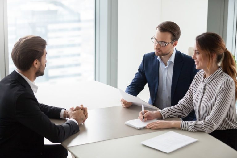 A man and woman taking notes and sitting across from a man at a table. They are all dressed in business wear.