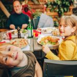 A family sits on a table as the younger children look towards the camera. On the table are burgers and pizza.