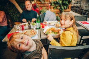 A family sits on a table as the younger children look towards the camera. On the table are burgers and pizza.