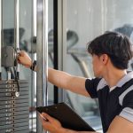 A close-up of a young man holding a clipboard while inspecting the pulley system of a workout machine.