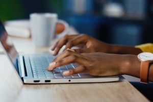 A close-up view shows a person wearing a brown leather watch and a yellow sweater typing on an open silver laptop.