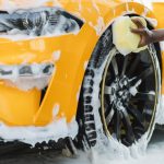 A person using a large, light yellow sponge to scrub suds againt the black wheel of a yellow-orange luxury sports car.