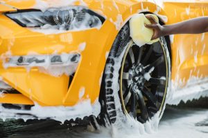 A person using a large, light yellow sponge to scrub suds againt the black wheel of a yellow-orange luxury sports car.