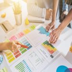 A meeting table with documents, charts, sticky notes, and a red pen, as hands point to a colored note sheet.
