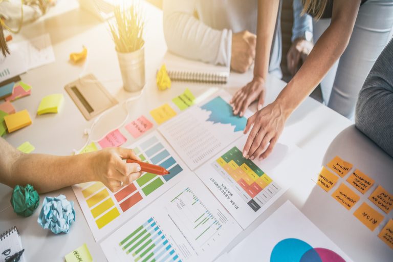 A meeting table with documents, charts, sticky notes, and a red pen, as hands point to a colored note sheet.