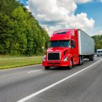 A red semi-truck traveling down a highway, flanked by lush green trees, with a smaller white truck following closely behind.
