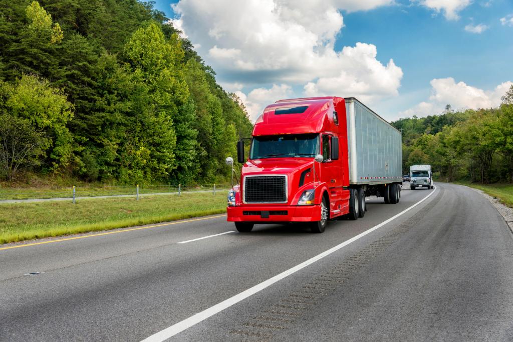 A red semi-truck traveling down a highway, flanked by lush green trees, with a smaller white truck following closely behind.