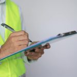 A person, dressed in a white button-up shirt and yellow vest, standing beside a wall and holding a blue clipboard and pen.