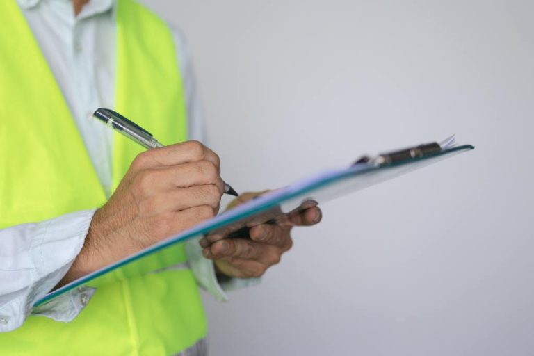 A person, dressed in a white button-up shirt and yellow vest, standing beside a wall and holding a blue clipboard and pen.
