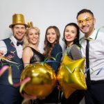 A group of event guests gathered together for a photo in a photo booth. They're smiling, wearing props like hats and glasses.