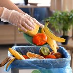 A person wears plastic kitchen gloves as they push food waste into the garbage bin. The plate has a pepper and a banana.