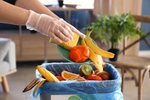 A person wears plastic kitchen gloves as they push food waste into the garbage bin. The plate has a pepper and a banana.