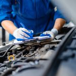 A close-up of an automotive mechanic doing maintenance and checking a list of things while wearing gloves.