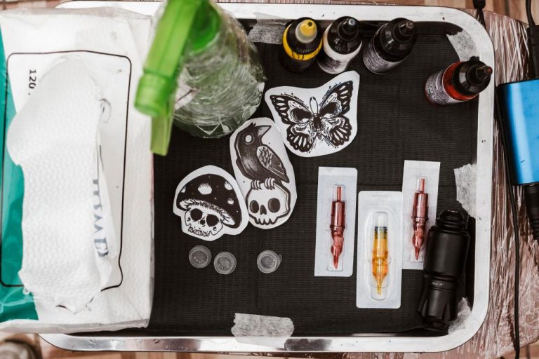 A tray with a black mesh mat holding various tattoo supplies, including stencils, ink, needles, a tattoo gun, and wipes.