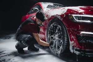 A man with a graying beard is washing down a red car. The car is covered in soap suds and the man is wearing overalls.