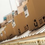 Six brown boxes on a conveyor belt moving on a production line inside a brightly lit industrial facility.