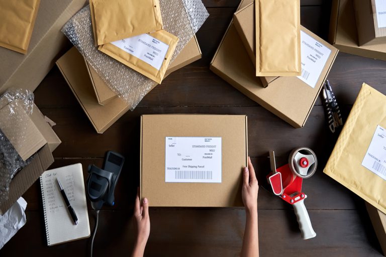 An overhead view of a person packing e-commerce shipping boxes on a wooden table, surrounded by padded envelopes.