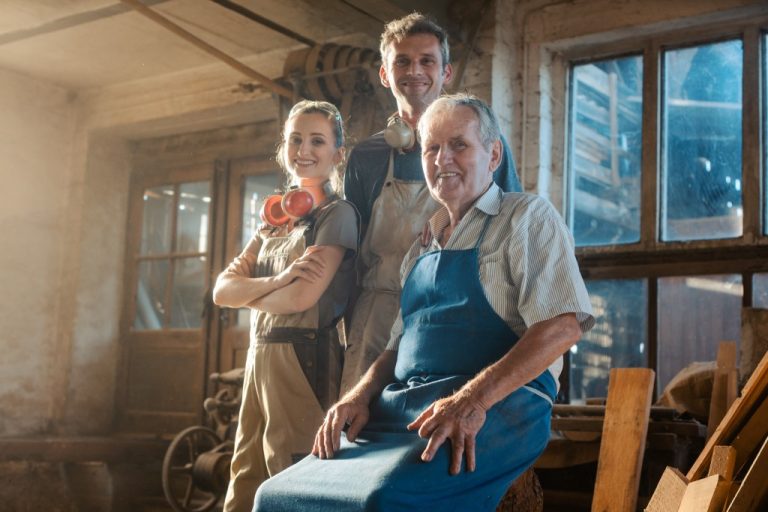 A group of three carpenters standing in their workshop in aprons. The oldest is sitting while the other two are standing.